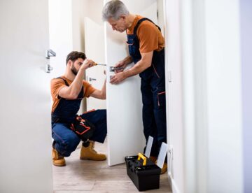 Full length shot of two locksmith, repairmen, workers in uniform installing, working with house door lock using screwdriver. Repair, door lock service.