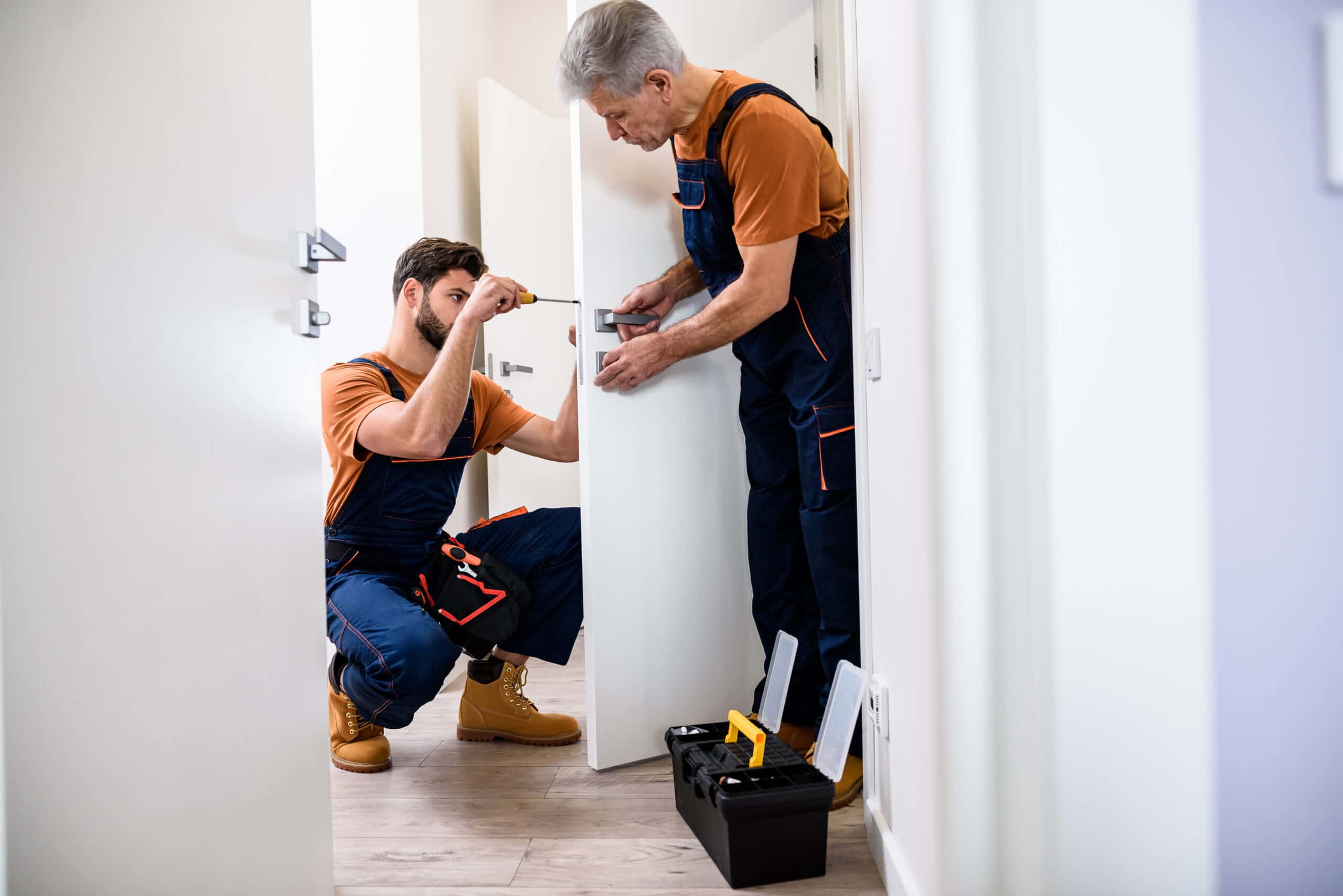 Full length shot of two locksmith, repairmen, workers in uniform installing, working with house door lock using screwdriver. Repair, door lock service.